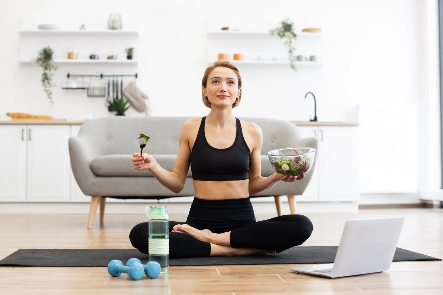 A woman sitting cross-legged at home with a bowl of salad, water and light workout gear beside her